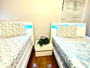 Una habitación pequeña y limpia con dos camas individuales, cada una con ropa de cama y almohadas estampadas con motivos florales. Hay una mesita de noche pequeña con una planta y algunos objetos encima. La habitación tiene un diseño sencillo y funcional, con suelo de madera y una ventana con persianas.