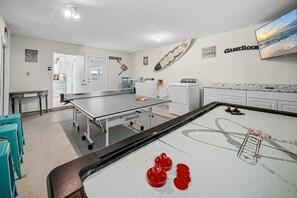 Una mesa de billar en una habitación con pared blanca, una tabla de surf colgada y una pantalla de televisión mostrando una escena de playa.