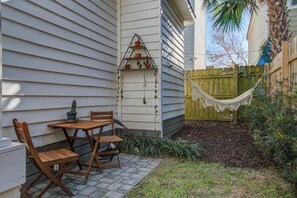 A patio with a wooden table and chairs, a hammock, and a tiered plant stand against a white house. - Historic Cottage • walk to King St, fenced yard • patio • hammock • string light (Charleston)