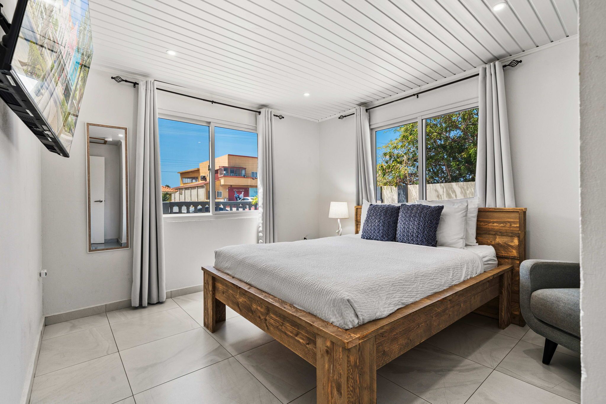 A bedroom with a wooden bed, a nightstand, a lamp, and a view of buildings through large windows.