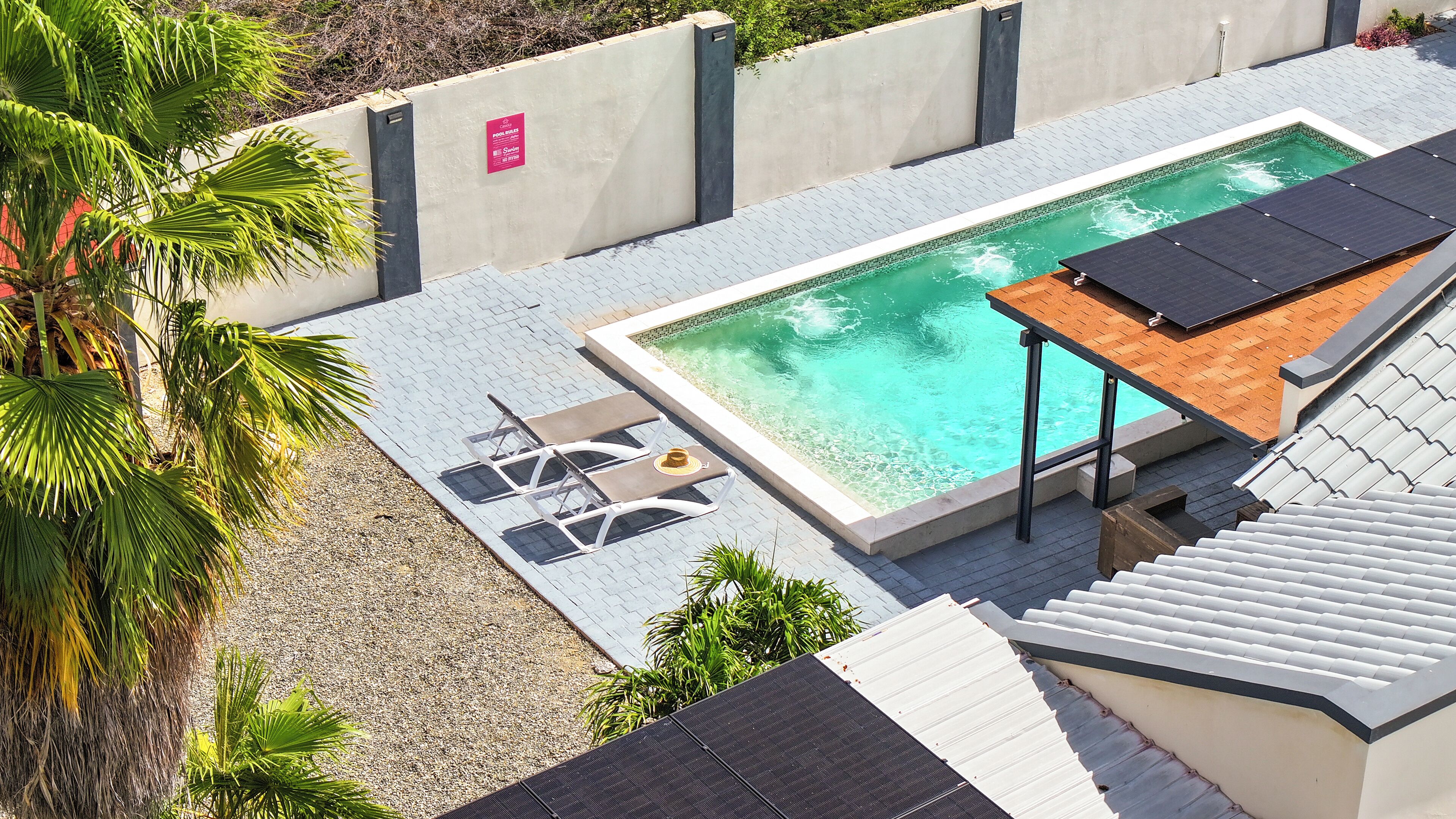 A rooftop pool with a wooden deck and lounge chairs, surrounded by palm trees and a concrete wall with a sign.