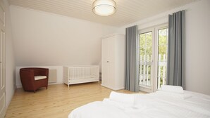 A bedroom with a large bed, a wooden floor, a white crib, a red chair, and a window with curtains. - Vacation home Wiking - in a quiet location, beautiful vacation home with terrace and garden (Binz)