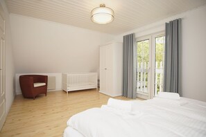 A bedroom with a large bed, a wooden floor, a white crib, a red chair, and a window with curtains.