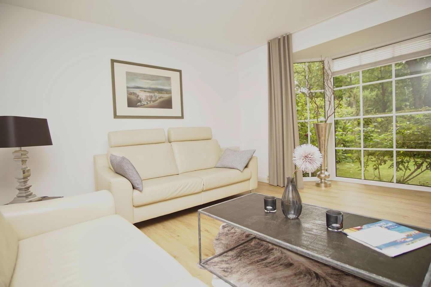 Un salon moderne avec un canapé beige, une table basse, une lampe et une grande fenêtre donnant sur un paysage verdoyant.