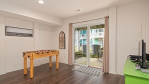 A room with a foosball table, a green desk with a computer and keyboard, and a sliding glass door leading to an outdoor area with a balcony.