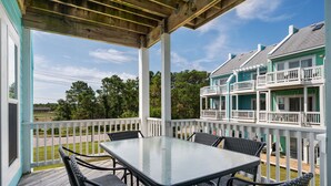 A covered outdoor area with a glass-top table and wicker chairs, surrounded by a white railing, overlooking a residential area with houses.
