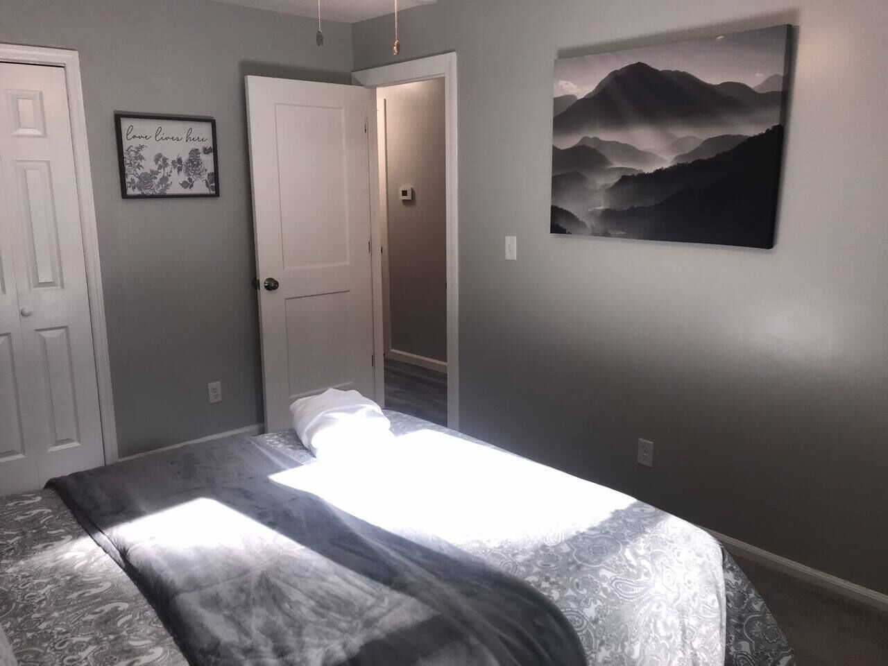 A bedroom with a bed, a door, a framed quote on the wall, and a landscape painting.