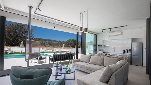 A modern living room with a large sofa, a coffee table, and a TV. There is a kitchen area with white cabinets and a refrigerator. A pool is visible outside the window. - Protaras Elite Pearl Villa EP14 -  a villa that sleeps 11 guests  in 5 bedrooms (Protaras)