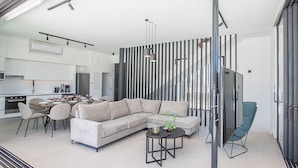 A modern living space with a sofa, dining table, and chairs, a kitchen area, and a large window. - Protaras Elite Pearl Villa EP14 -  a villa that sleeps 11 guests  in 5 bedrooms (Protaras)