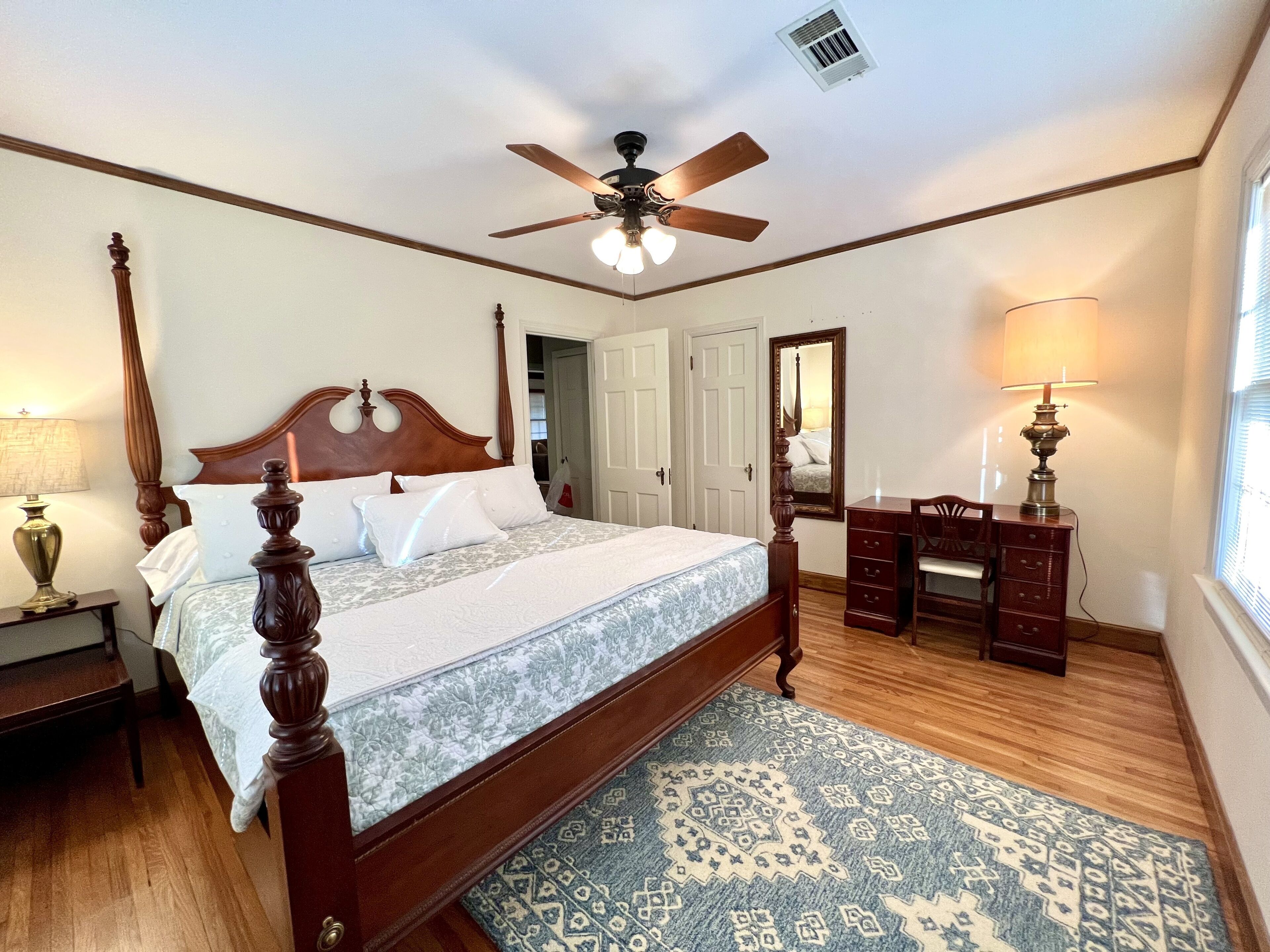A bedroom with a four-poster bed, a ceiling fan, a dresser, a mirror, and a window with blinds.