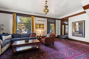 Une chambre d’hôtel avec un canapé, un fauteuil et une table basse. Il y a une grande fenêtre avec des rideaux, un tableau au mur et une affiche.