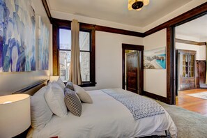 A bedroom with a large bed, a painting on the wall, and a view of the outside through a window.