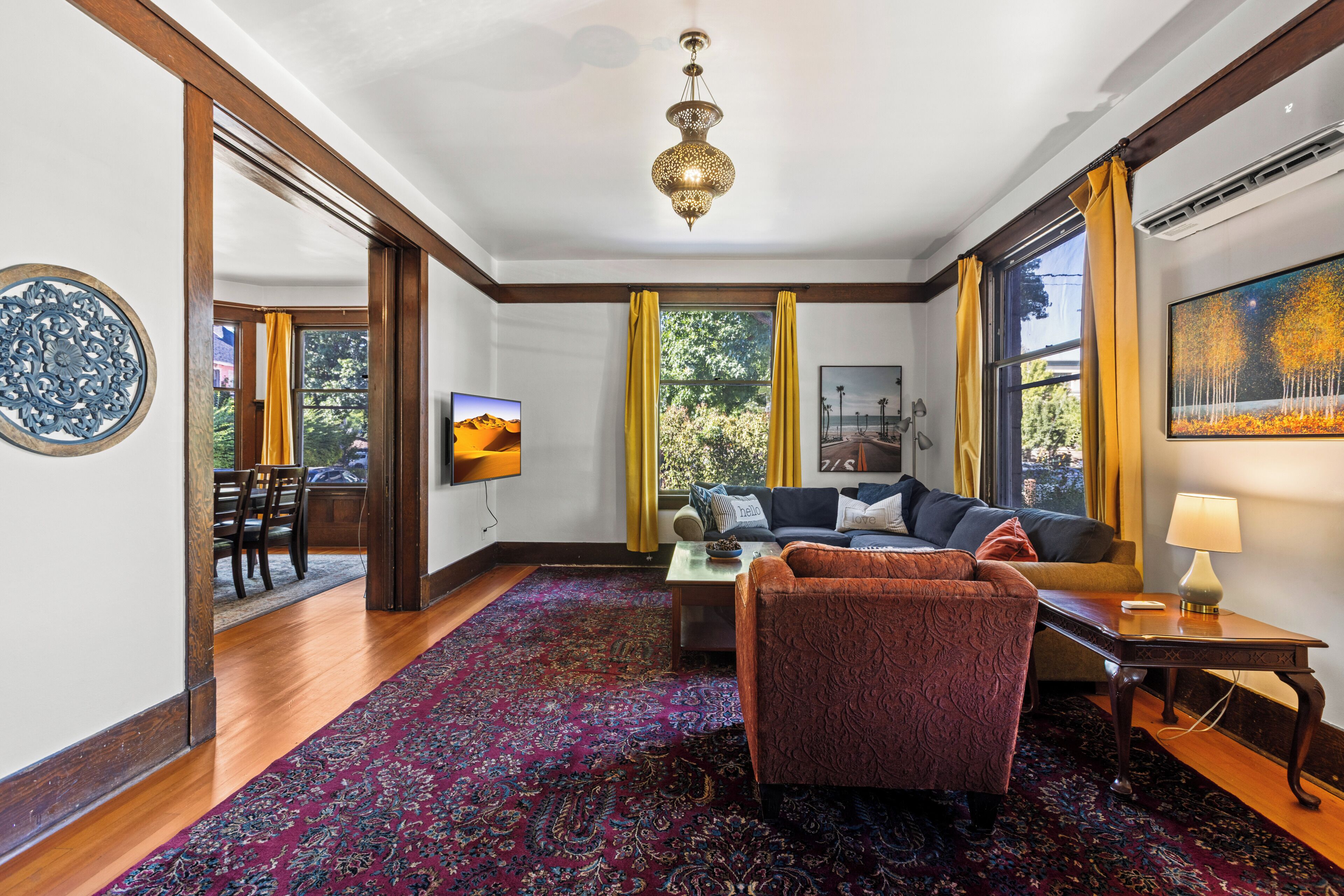 A well-lit living room with a sofa, armchair, and a TV. There is a large window with curtains, a patterned rug, and a chandelier.