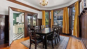 Uma sala de jantar com uma mesa de madeira escura, seis cadeiras, um lustre e grandes janelas com cortinas amarelas.