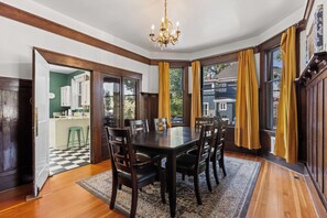 A dining room with a dark wooden table, six chairs, a chandelier, and large windows with yellow curtains. - Art House Apt 1 Northend Historic Apartment (Boise)