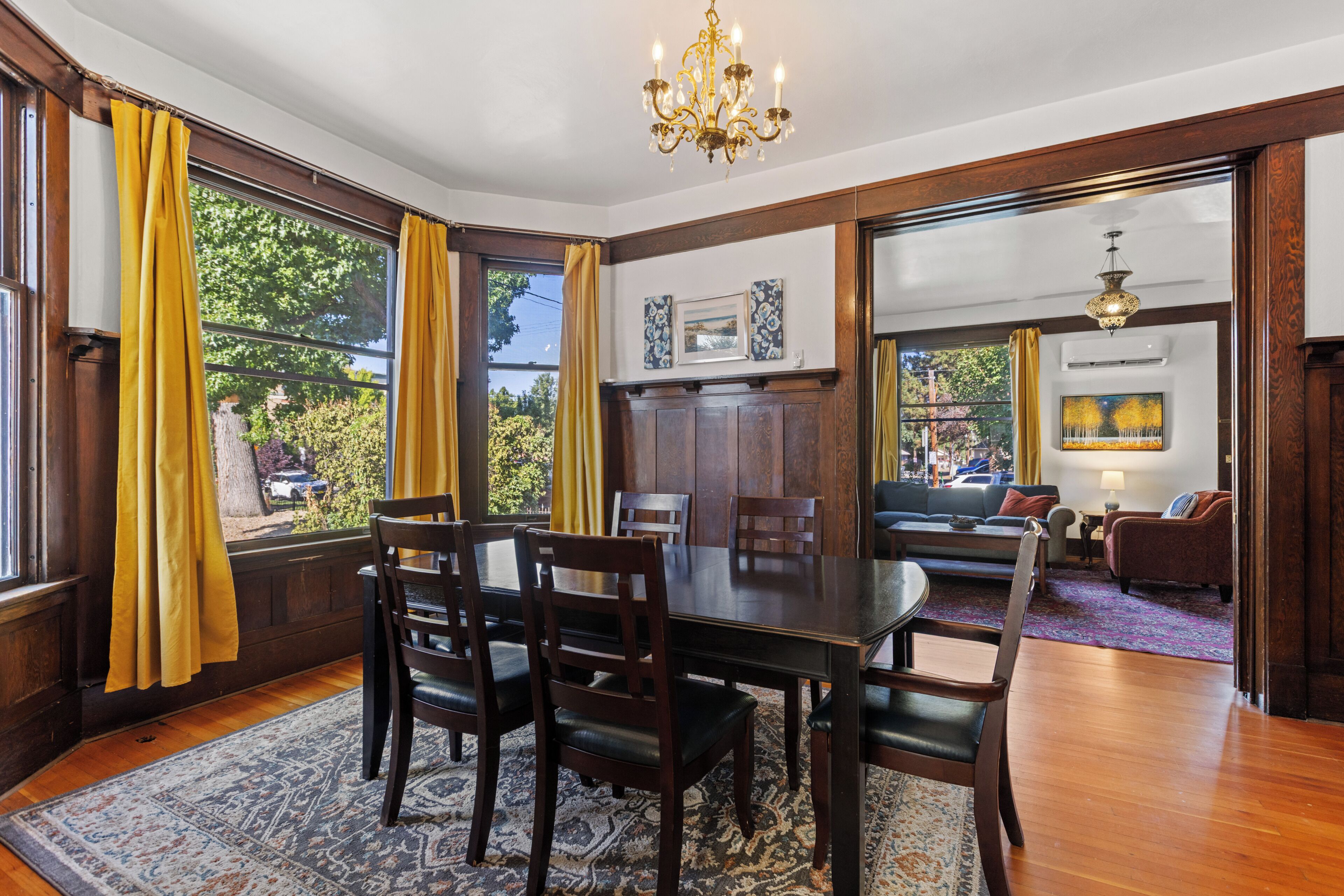 A dining room with a wooden table and chairs, a chandelier, and large windows with yellow curtains.