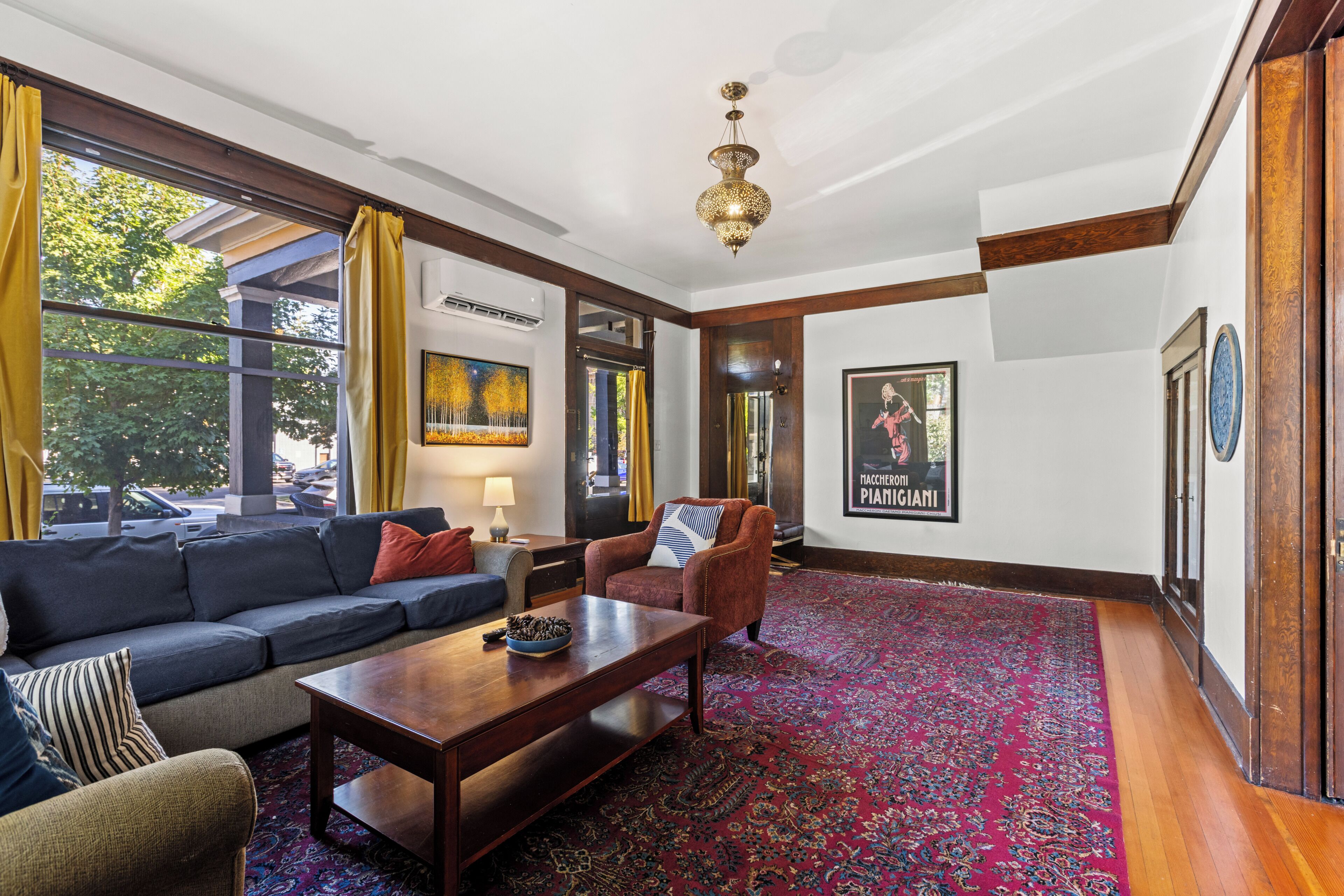 A room with a dark brown sofa, a wooden coffee table, and a patterned rug. There is a large window with yellow curtains, and a poster on the wall.