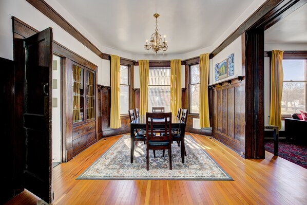 A dining room with a wooden table and chairs, a chandelier, and large windows with curtains.