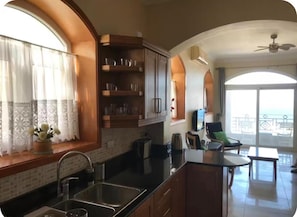 A kitchen with a sink, cabinets, and a window with curtains. There is a balcony with a view of the sea.