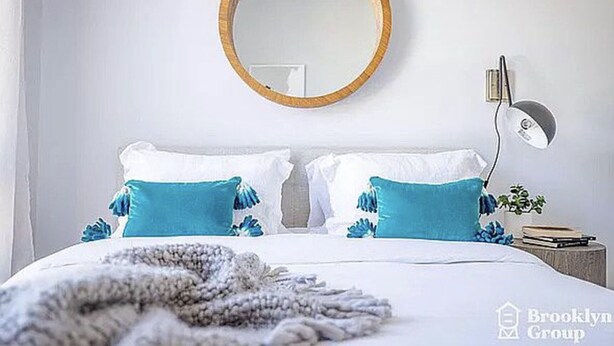 Ein Schlafzimmer mit einem runden Spiegel, zwei weißen Kissen, einer blauen Tagesdecke und einer Nachttischlampe.