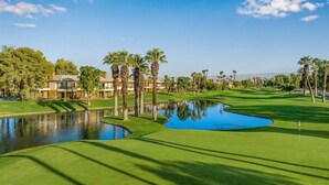 A golf course with a pond, palm trees, and a building complex in the background. - Palm Desert | Desert Oasis | Golf & Resort Pool (Palm Desert)