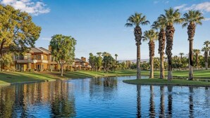 A golf course with a pond, palm trees, and a building with multiple balconies. - Palm Desert | Desert Oasis | Golf & Resort Pool (Palm Desert)