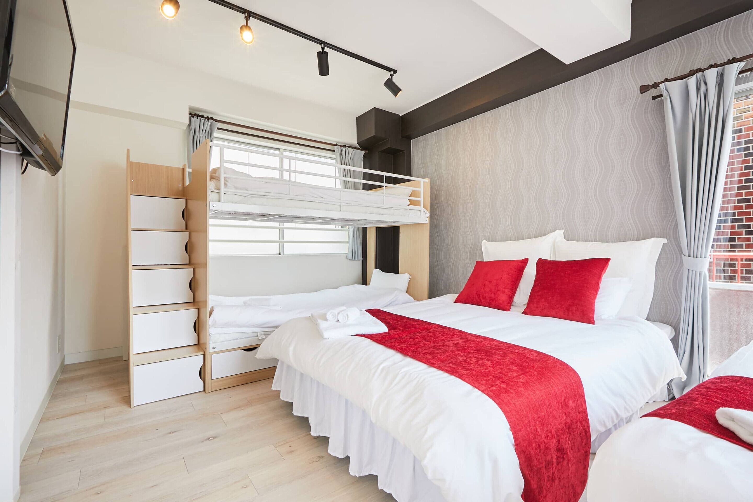 A modern hotel room with a bunk bed, a wooden wardrobe, a large bed with red accents, and a window with curtains.