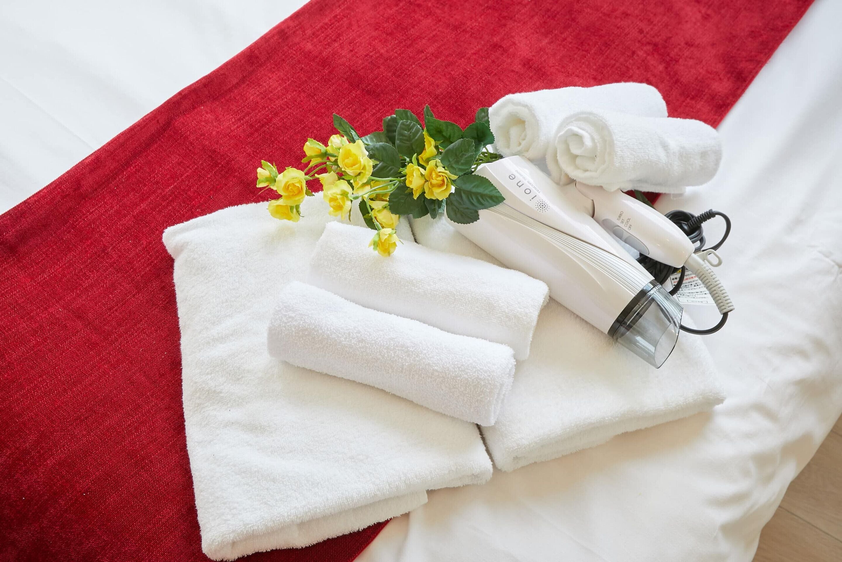 A neatly arranged set of white towels, a hairdryer, and a small bouquet of yellow flowers on a bed with a red blanket.