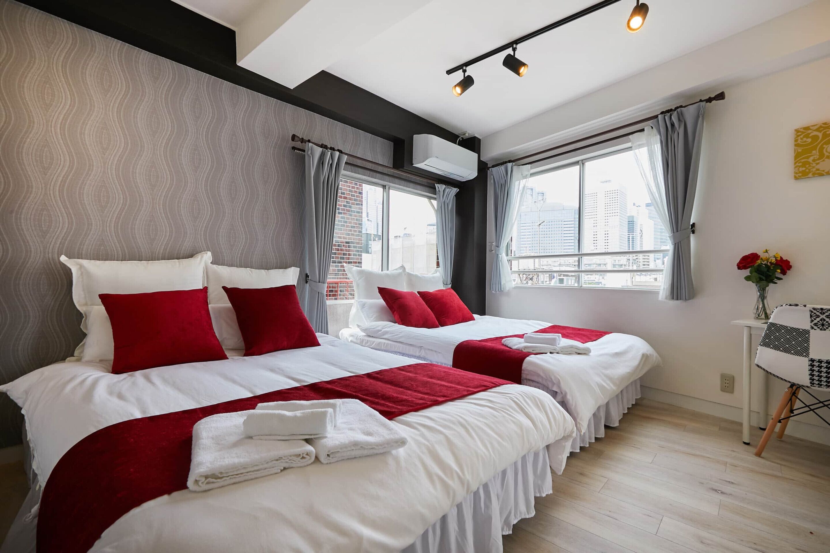A hotel room with two beds, a small table with a chair, a window with curtains, and an air conditioning unit.