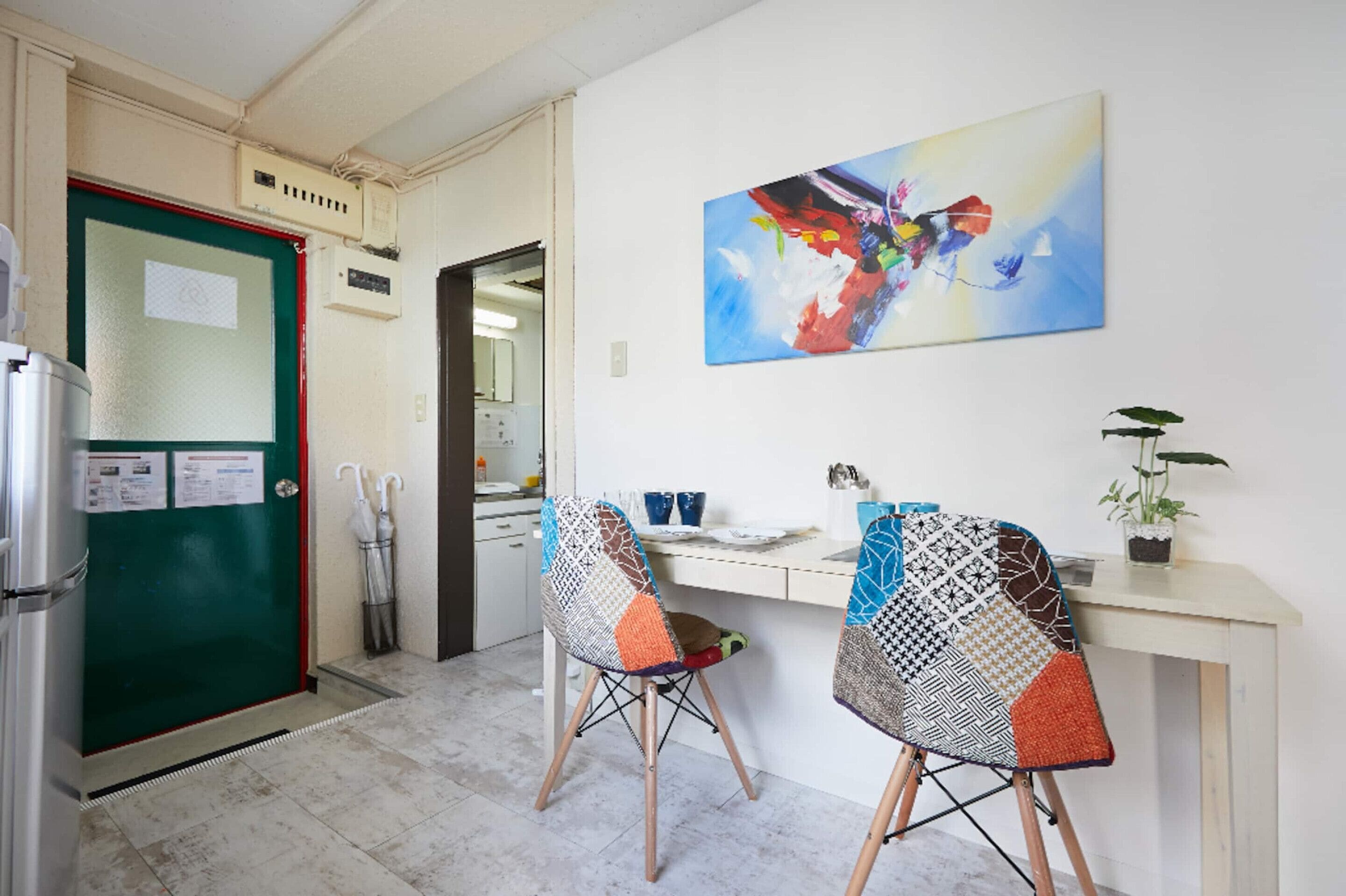 A kitchen with a refrigerator, a green door, a white countertop, two chairs, a table, a painting, and a potted plant.