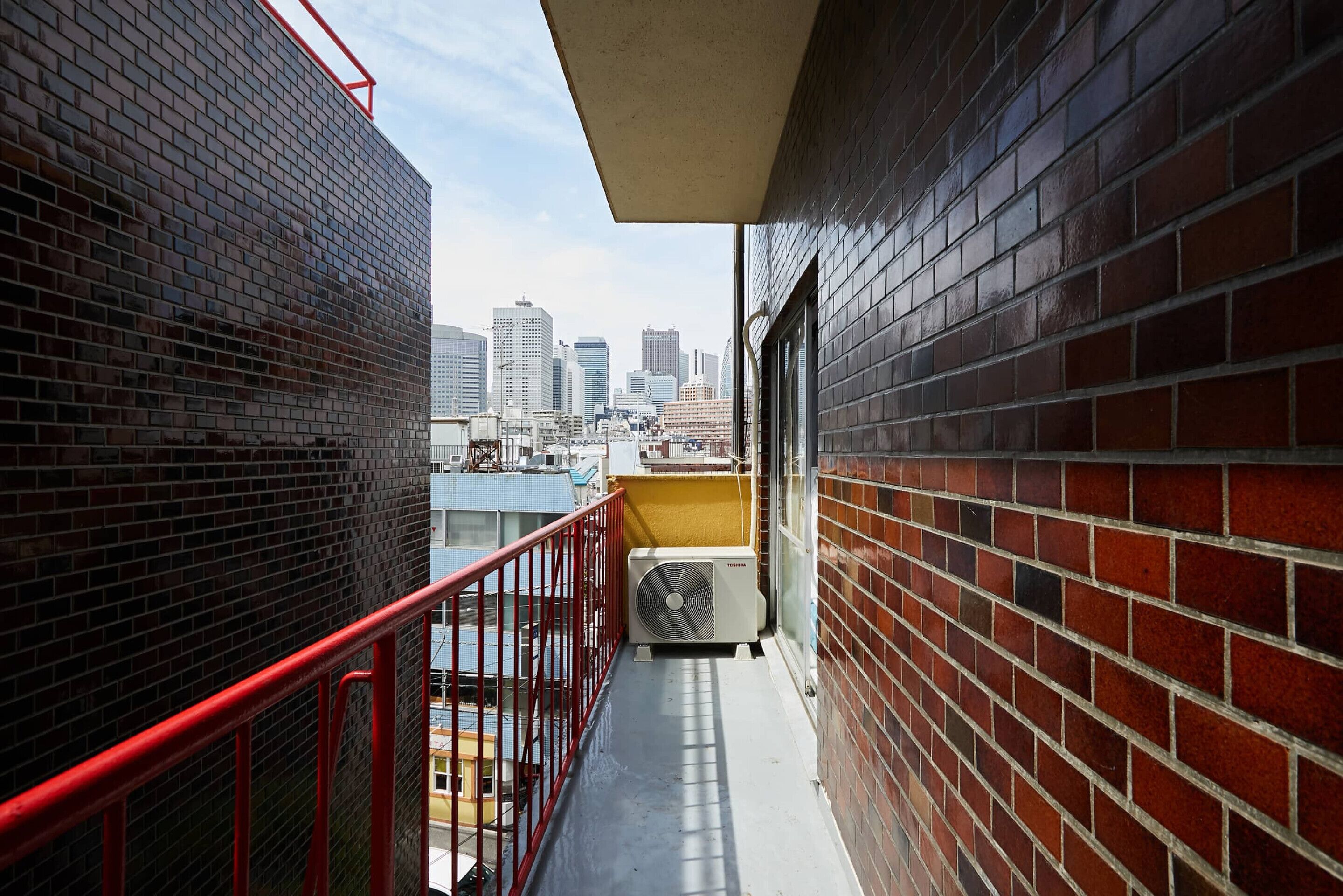 Ein schmaler Balkon mit roten Geländern, einer Ziegelwand und Blick auf die Skyline der Stadt.