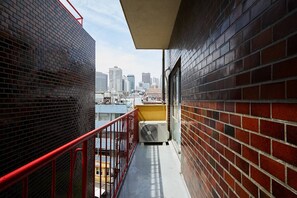 A narrow balcony with red railings, a brick wall, and a view of the city skyline.