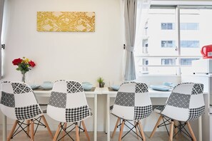 A dining area with four chairs, a table, a vase with flowers, a wall-mounted artwork, and a window with a view of buildings.