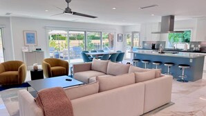 A modern living room with a large beige sectional sofa, a coffee table, and a TV. There is a dining area with blue chairs and a kitchen with a bar. - Fairway Luxe: New Remodel on Indian Canyons Course (Palm Springs)
