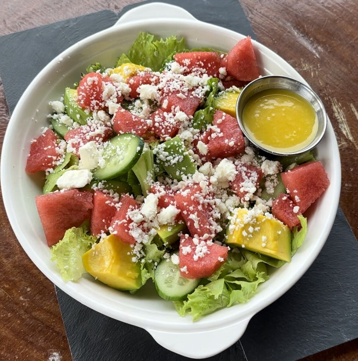 Un tazón de ensalada con sandía, aguacate, queso feta y un recipiente pequeño con aderezo.