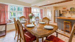 A dining area with a wooden table set for four, surrounded by chairs with red cushions. A vase with flowers is on the table, and there is a view of a kitchen with a refrigerator and various kitchen items. - Woodmancote Manor Cottage, near Cirencester - sleeps 6 guests in 3 bedrooms (Cirencester)