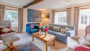 A cozy living room with a blue sofa, a grey armchair, and a wooden coffee table. There is a colorful abstract painting on the wall and a window with curtains. - Woodmancote Manor Cottage, near Cirencester - sleeps 6 guests in 3 bedrooms (Cirencester)