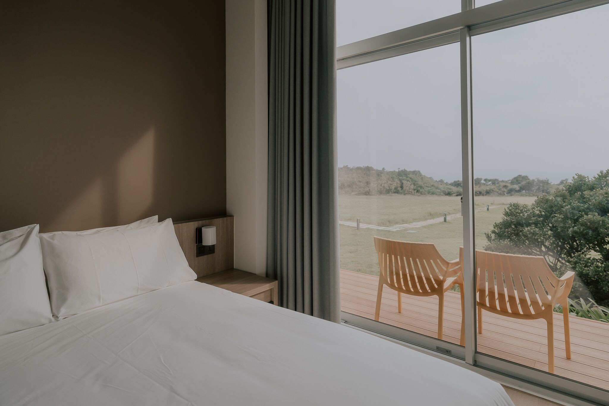 A bedroom with a large bed, a wooden nightstand, and a view of a deck with chairs.