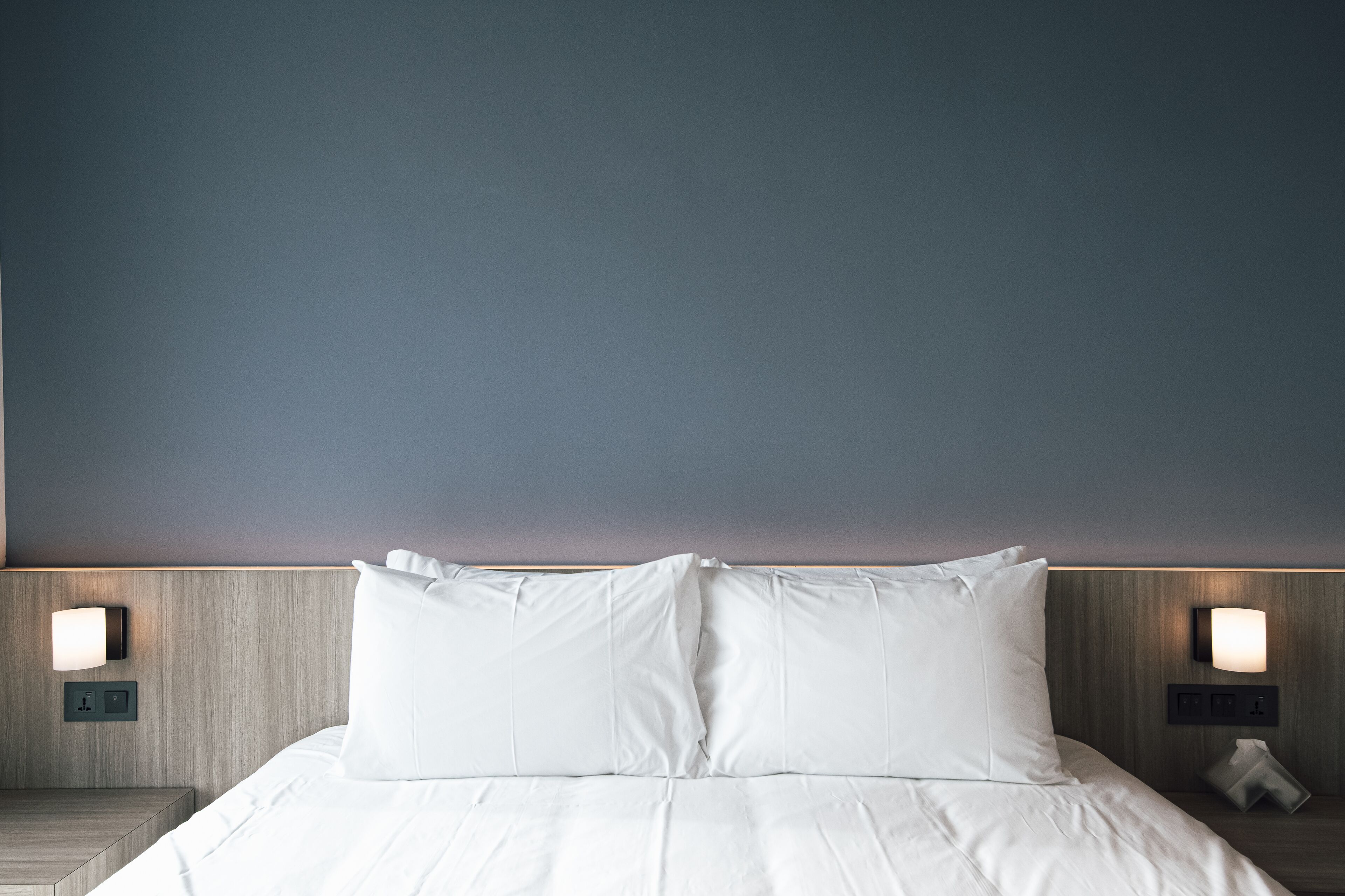 A neatly made bed with white linens and pillows, a wooden headboard, and two wall-mounted nightstands with lamps and a telephone.
