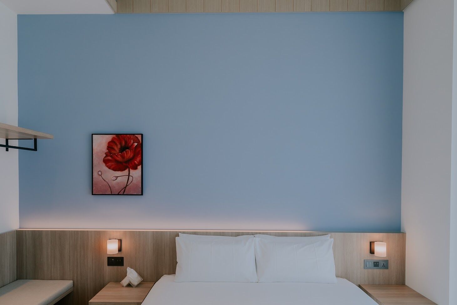 A hotel room with a bed, two bedside tables, a nightstand, a wall-mounted light, and a framed picture of a red flower.