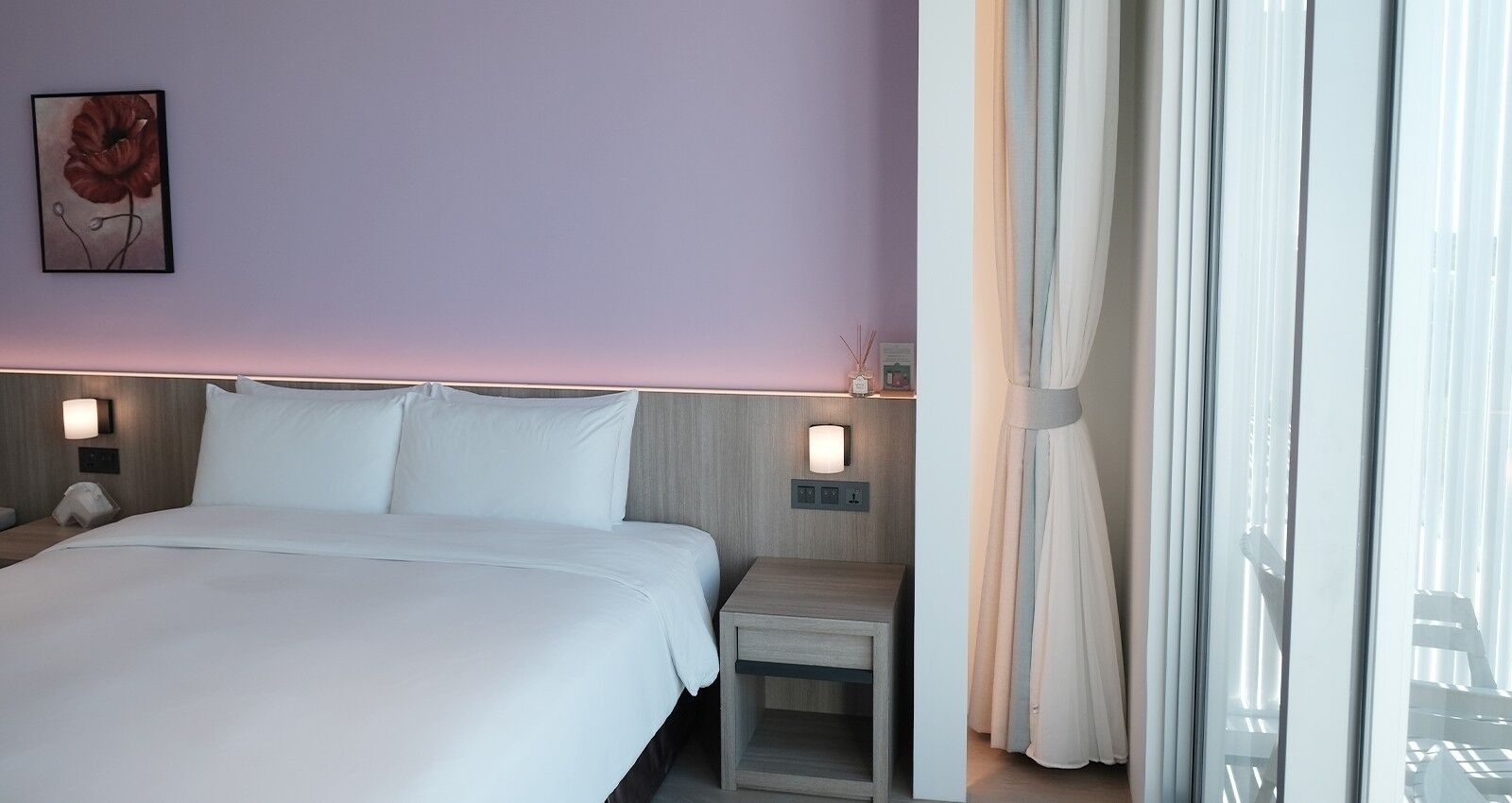 A hotel room with a large bed, bedside tables with lamps, a wall-mounted artwork, and a view through sheer curtains.