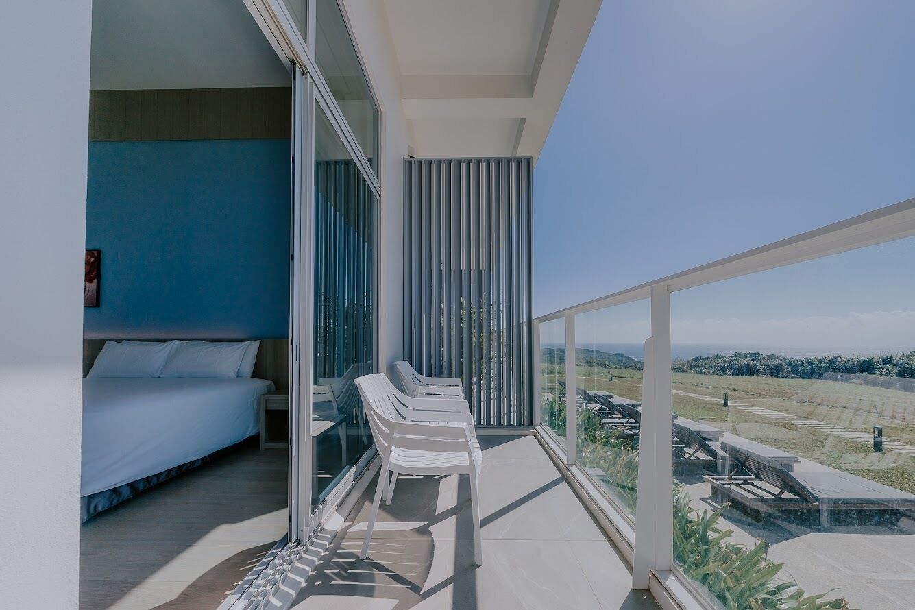 A balcony with white chairs, a bed with white bedding, and a view of a landscaped area.