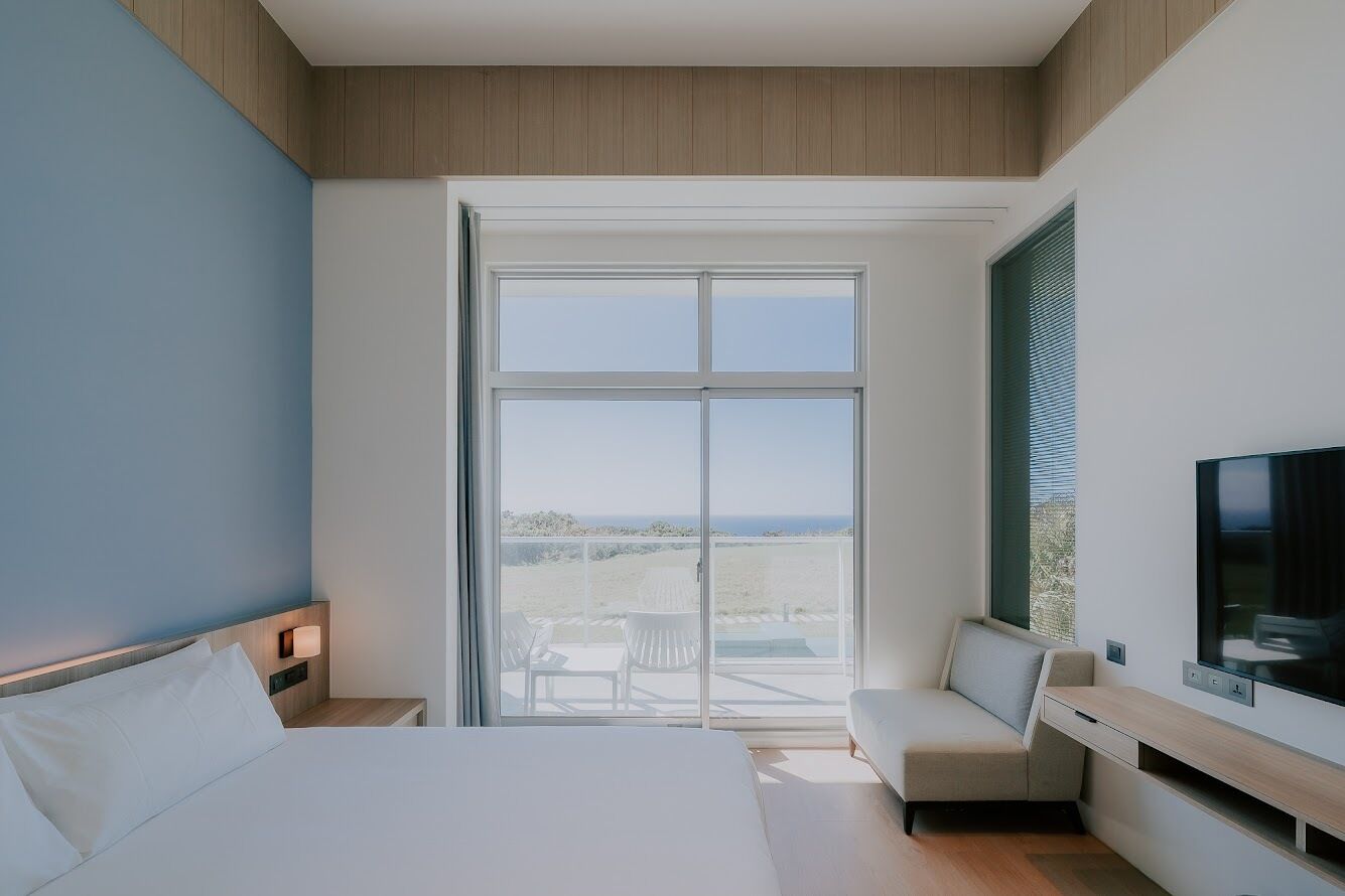 A modern hotel room with a large bed, a seating area, a TV, and a balcony with a view.