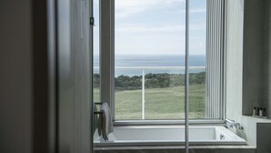 Et badeværelse med et stort vindue, der giver udsigt over havet og et græslandskab.