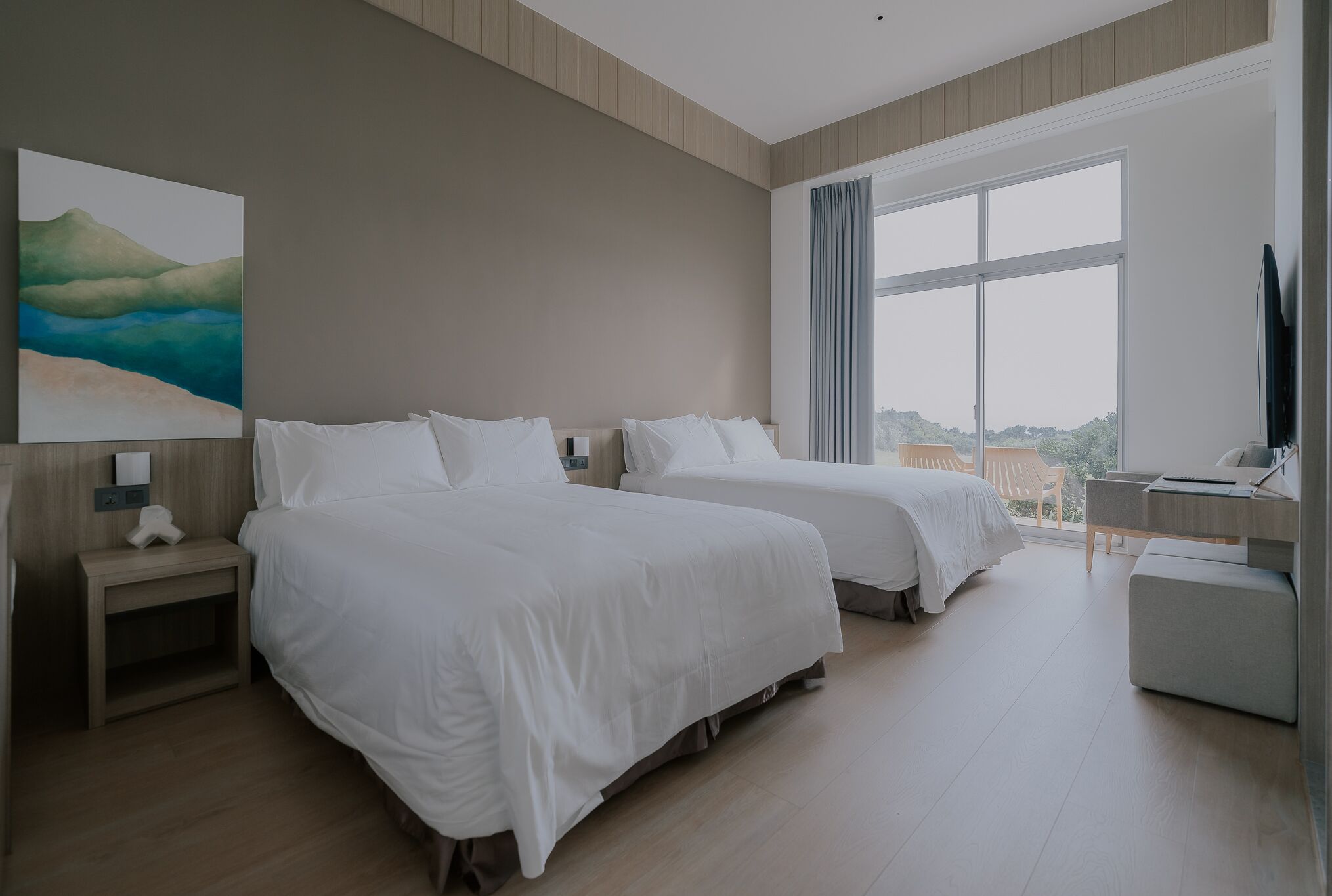 A hotel room with two beds, a desk, a chair, a large window, and a painting on the wall.