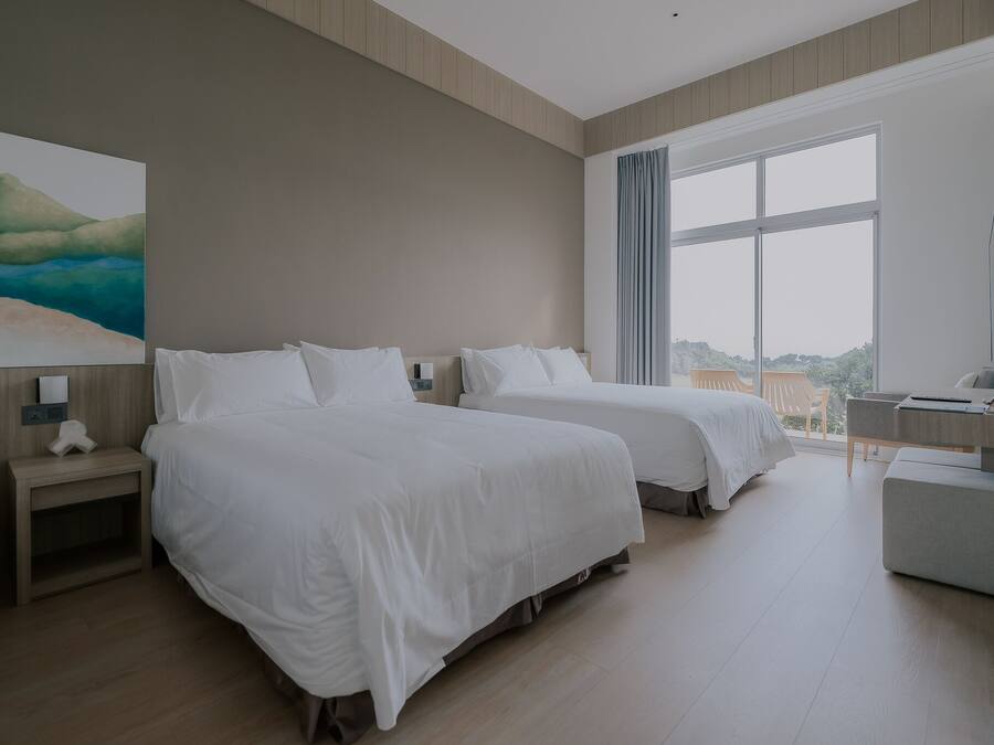 A hotel room with two beds, a desk, a chair, a large window, and a painting on the wall.