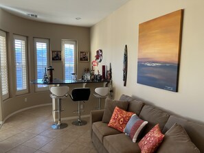 Un salon avec un canapé marron, deux tabourets de bar blancs, un coin bar et un grand tableau accroché au mur.