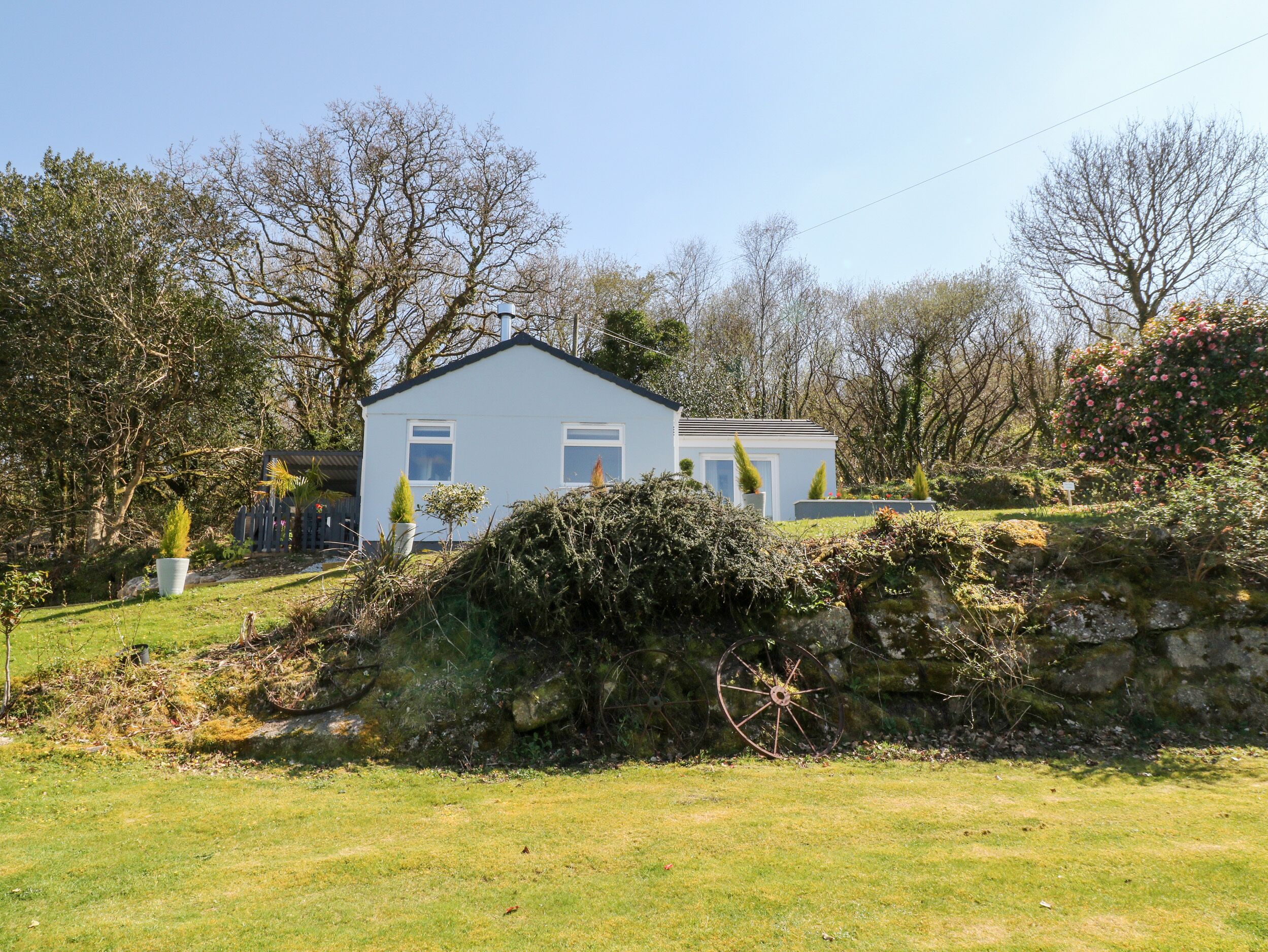 A small, single-story house with a white exterior and a dark roof, surrounded by a well-maintained garden with various plants and a stone wall.