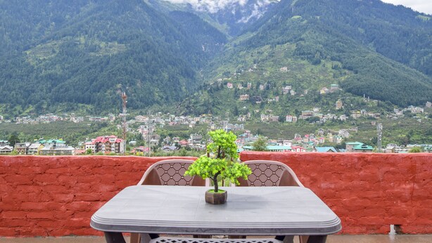 A table with a potted plant in front of a red wall, overlooking a mountainous landscape with buildings in the distance. - Madhuvan 2BHK, W/ Food, parking, Mountain View (Manali)