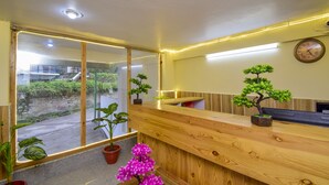 A reception area with wooden reception desk, potted plants, a clock on the wall, and a view of the outside through large windows. - Madhuvan 2BHK, W/ Food, parking, Mountain View (Manali)